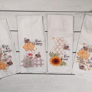 Pode incluir: Conjunto de quatro toalhas de cozinha brancas com designs de abelhas e citações inspiradoras. As toalhas apresentam abelhas, favos de mel, girassóis e elementos florais. As citações são "Bee the Change", "Bee Happy", "Bee Kind", e "Home Sweet Hive".