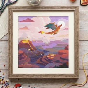 May include: A framed cross-stitch embroidery featuring a pixelated orange dragon flying over a volcanic landscape with purple clouds and a full moon. The artwork is displayed in a light brown wooden frame with a white mat, surrounded by craft supplies.