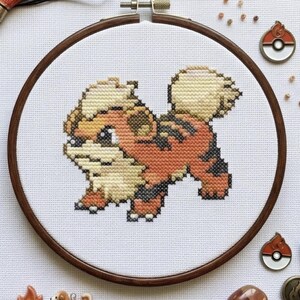 May include: A cross-stitch embroidery project in a circular wooden hoop featuring a pixelated orange and cream-colored creature with black stripes. The design is centered on white fabric, accompanied by orange embroidery floss and gold scissors.