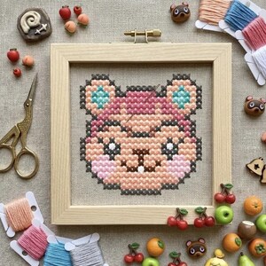 Può includere: Un telaio da ricamo quadrato in legno contenente un motivo a punto croce di un volto animale stilizzato con filo rosa, pesca e verde acqua. Il disegno presenta un ago inserito nel tessuto, circondato da fili da ricamo, forbici e ciondoli.