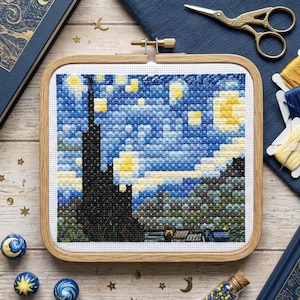 May include: A square wooden embroidery hoop containing a cross-stitch pattern depicting a night sky with a dark cypress tree, yellow stars, and a crescent moon. The design features shades of blue, yellow, and green thread on white fabric.
