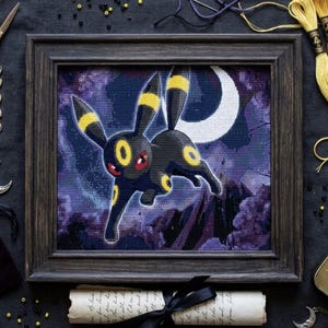 May include: A framed cross-stitch embroidery featuring a black creature with yellow rings and red eyes leaping against a purple night sky with a white crescent moon. The artwork is displayed in a dark wooden frame with a visible grid texture.