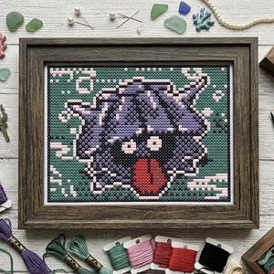 May include: A cross-stitch embroidery in a dark wood frame featuring a purple, horned creature with a wide open mouth and red tongue against a dark green background. The piece is surrounded by embroidery floss, scissors, and small decorative items.