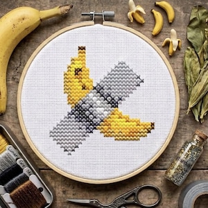 Puede incluir: Un bastidor de bordado circular de madera que contiene un patrón de punto de cruz de una banana amarilla pegada a un fondo blanco con cinta adhesiva plateada. El diseño presenta puntadas de hilo amarillo, gris y blanco sobre tela.