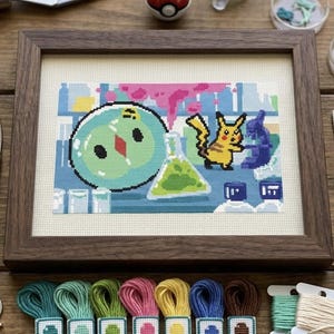 May include: A framed cross-stitch embroidery featuring a pixelated scene with a round green creature, a yellow Pikachu character, and a laboratory flask with green liquid. The artwork is displayed in a dark wood frame on a wooden surface.