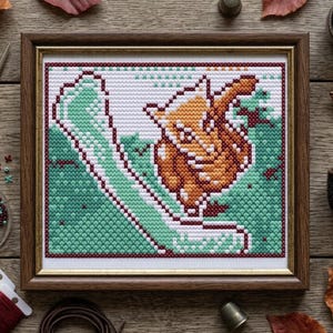 May include: A framed cross-stitch embroidery piece featuring a stylized orange fox curled up against a background of green and white geometric patterns. The artwork is enclosed in a dark brown wooden frame with a thin gold inner border.
