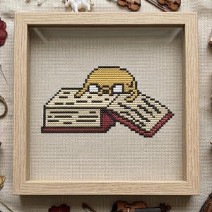 May include: A square light wood frame containing a cross-stitch embroidery on beige fabric. The design features a yellow cartoon character with white eyes lying across an open book with white pages and a dark red cover, rendered in pixelated stitches.
