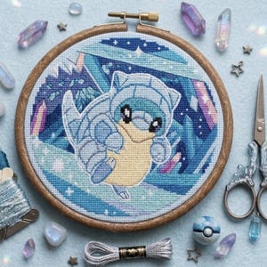 May include: A circular wooden embroidery hoop containing a cross-stitch pattern of a blue and cream-coloured creature with a shell, surrounded by blue geometric shapes and crystals. Includes blue embroidery floss and metal scissors.