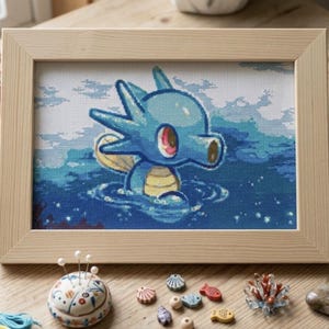 May include: A cross-stitch embroidery in a light wood frame featuring a blue, seahorse-like creature with a yellow belly and red eye, partially submerged in blue water. The design includes white highlights and ripples, presented in a rectangular format.