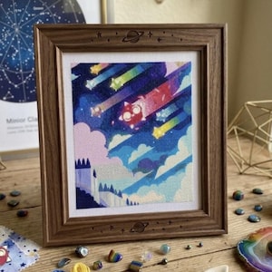 May include: A framed cross-stitch embroidery featuring a stylized red cat-like creature flying through a night sky with shooting stars, clouds, and a castle silhouette. The wooden frame is engraved with small planet and star motifs at the top and bottom.
