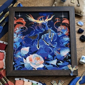 May include: A framed cross-stitch embroidery featuring a stylized deer with glowing antlers amidst dark blue floral patterns and stars. The piece is surrounded by embroidery floss skeins, needles, a small bottle, a key, and gold scissors.