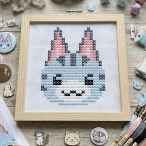 May include: A cross-stitch embroidery of a stylised light blue cat face with pink-tipped ears, dark grey stripes, and a white muzzle, framed in a light wood square frame, surrounded by various cat-themed pins, embroidery floss, and a paper pattern.