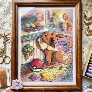 Puede incluir: Un bordado en punto de cruz enmarcado que representa a un personaje Eevee marrón de pie en una habitación con una bola roja y blanca, un objeto en forma de estrella y una forma morada en el suelo. La escena incluye una mesa y una planta.