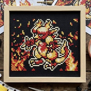 May include: A square wooden frame containing a cross-stitch embroidery on black fabric. The design features a pixelated creature in shades of red, orange, and yellow, surrounded by scattered flame-like shapes in the same vibrant color palette.