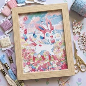 Puede incluir: Un bordado en punto de cruz enmarcado que muestra a un personaje Sylveon blanco en un campo de flores rosas y rojas bajo un cielo azul, rodeado de bobinas de hilo, un rollo de encaje, pines esmaltados y tijeras de cigüeña doradas.
