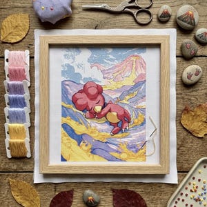 May include: A cross-stitch project in a light wood frame featuring a red creature sleeping on a yellow and purple landscape with a volcano in the background. The set includes a pincushion, embroidery scissors, thread bobbins, and painted stones.