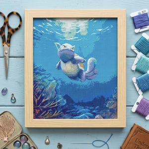 May include: A framed cross-stitch embroidery depicting a light blue aquatic creature swimming underwater among coral reefs. The artwork is surrounded by sewing supplies including metal scissors, colorful embroidery thread spools, and small charms.