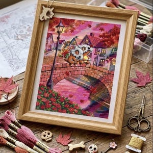 May include: A framed cross-stitch embroidery depicting two creatures with skull-like masks sitting on a stone bridge in a town at sunset. The scene features pink and orange hues, a tall street lamp, and houses. A small skull charm hangs from the frame.