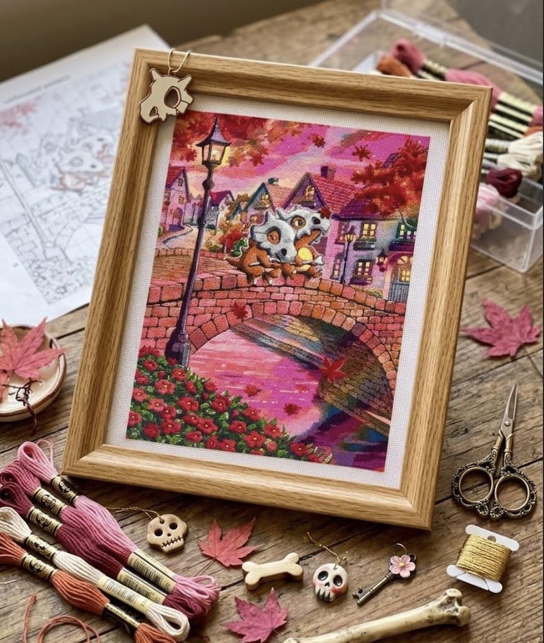 May include: A framed cross-stitch embroidery depicting two creatures with skull-like masks sitting on a stone bridge in a town at sunset. The scene features pink and orange hues, a tall street lamp, and houses. A small skull charm hangs from the frame.