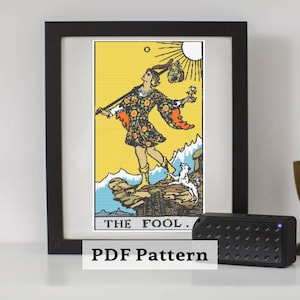 May include: Framed cross-stitch of The Fool tarot card. The image features a figure in patterned clothing, a dog, and a sun. The background is yellow, with blue and white accents. The text "THE FOOL" is at the bottom.