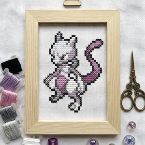 May include: A cross-stitch embroidery of a Mewtwo character in shades of purple, white, and gray, framed in a light wood frame. Beside the frame are several spools of embroidery thread, a pair of vintage-style metal scissors, and small beads.