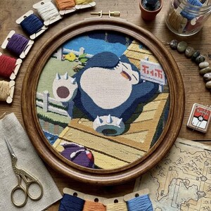 Könnte beinhalten: Ein runder hölzerner Stickrahmen mit einem Kreuzstichbild, das ein großes blaues und cremefarbenes Snorlax-Wesen zeigt, das auf einem gelben Holzsteg liegt. Die Szene umfasst einen weißen Zaun, grünes Gras und ein kleines violettes Wesen.