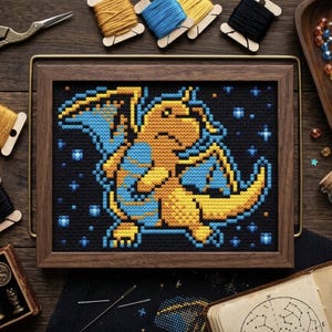 Op de afbeelding: Een ingelijst kruissteekborduurwerk met een gepixelde gele en blauwe draakachtige figuur op een zwarte achtergrond. Het ontwerp bevat kleine blauwe en gele stervormige accenten rondom de centrale figuur, alles gevat in een rechthoekige houten lijst.