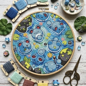 May include: A circular wooden embroidery hoop containing a cross-stitch pattern of blue cartoon creatures swimming in water with lily pads and rocks. The scene is surrounded by embroidery floss on wooden spools, small decorative figurines, and scissors.