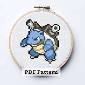 Puede incluir: Un patrón de punto de cruz de una criatura azul pixelada con forma de tortuga y caparazón beige, dentro de un bastidor de bordado de madera. El texto "PDF Pattern" se muestra en la parte inferior.