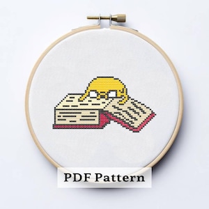 May include: Cross-stitch embroidery hoop featuring a yellow cartoon character resting on an open book. The book has beige pages and a red spine. The text "PDF Pattern" is at the bottom. The hoop is made of wood and has a brass adjustment screw.