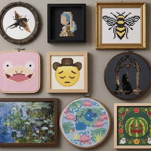 May include: A collection of eight framed cross-stitch artworks featuring diverse designs including a beetle, a pixelated portrait, a bee, a pink frog face, a cowboy emoji, a black cat in a window, a water lily pond, and a pumpkin.