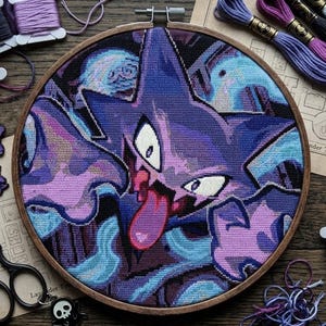 May include: A circular wooden embroidery hoop containing a cross-stitch design of a purple Gengar character with a long pink tongue, surrounded by blue and dark purple abstract shapes, accompanied by embroidery floss, scissors, and pins.