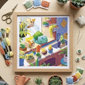 May include: A framed cross-stitch embroidery depicting a kitchen scene with a green creature, various potted plants, and a bird. The artwork is surrounded by embroidery floss, scissors, needles, and small succulent-themed buttons.