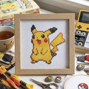 May include: A square wooden frame containing a cross-stitch embroidery of the yellow character Pikachu on white Aida fabric. The character features black-tipped ears, red cheeks, and a lightning bolt-shaped tail, rendered in a pixelated style.