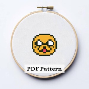 May include: A cross-stitch pattern of a cartoon character's face, featuring yellow, black, and red pixels, is centered on white fabric within a wooden embroidery hoop. The text "PDF Pattern" is displayed below the design.