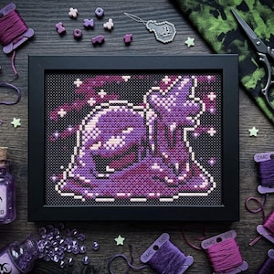 May include: A framed cross-stitch embroidery featuring a purple, blob-like creature with a star-shaped appendage against a black dotted background. The piece is surrounded by purple embroidery floss, small beads, and a glass bottle labeled JUK-ACID.