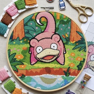 May include: A circular wooden embroidery hoop containing a cross-stitch project of a pink creature with a curled tail, large eyes, and a wide mouth, set against a green forest and blue water background, surrounded by colourful embroidery floss bobbins.