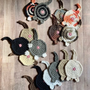 May include: A collection of crocheted cat butt coasters in various colors, including black, white, brown, gray, and orange. The coasters are made with a soft yarn and have a cute, whimsical design.