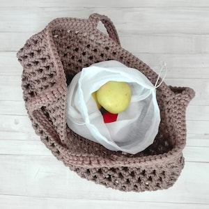 Könnte beinhalten: Eine handgefertigte, offen gewebte, taupefarbene Häkeltasche mit zwei Griffen. Im Inneren der Tasche befindet sich ein weißer Netz-Gemüsebeutel mit einem gelben Apfel und einer roten Paprika. Die Tasche ist zum Tragen von Lebensmitteln oder anderen Gegenständen bestimmt.