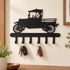 May include: A matte black metal wall-mounted key holder featuring a silhouette of a vintage pickup truck. The horizontal bar includes six downward-facing hooks for hanging items. Two sets of keys are currently hanging from the hooks on the rack.