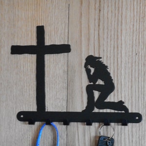 Puede incluir: Llavero de metal negro con una cruz y una figura arrodillada en silueta. El llavero tiene cuatro ganchos y está montado sobre una superficie de madera clara. Un cordón azul y llaves cuelgan de los ganchos.