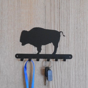 May include: Black metal key holder featuring a bison silhouette above a row of hooks. A blue lanyard and a set of keys hang from the hooks. The key holder is mounted on a light wood-grain surface.