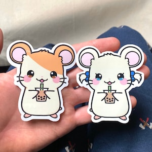 May include: Two cartoon hamster stickers holding cups of boba tea. The hamster on the left has brown and white fur and the hamster on the right has white fur. Both hamsters have pink ears and blue eyes.