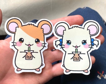 Könnte beinhalten: Zwei Cartoon-Hamster-Aufkleber, die Tassen mit Boba-Tee halten. Der Hamster links hat braunes und weißes Fell und der Hamster rechts hat weißes Fell. Beide Hamster haben rosa Ohren und blaue Augen.