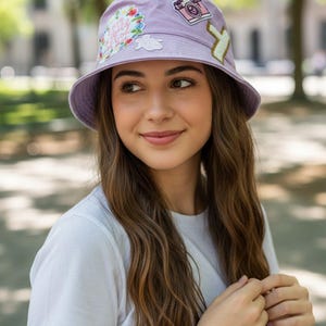 Peut inclure: Un chapeau bob lavande orné d'écussons brodés, dont un appareil photo, un motif floral et du texte. Le chapeau est porté par une personne portant un t-shirt blanc. L'arrière-plan est flou avec des arbres verts.