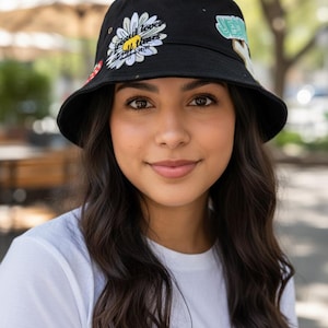 May include: Black bucket hat with a daisy and text design. The hat features a white daisy with yellow center and the words "lover all times". Additional patches include a red mushroom and a green design.