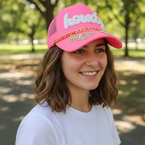 Puede incluir: Gorra de camionero rosa brillante con la palabra "howdy" en escritura blanca. Tiene un detalle de cadena dorada y la frase "love them anyway". La gorra tiene una parte trasera de malla y una visera curva.