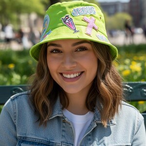 Puede incluir: Un sombrero de pescador verde lima adornado con parches coloridos, incluyendo la palabra "JESUS" y una cruz rosa. El sombrero se usa con una chaqueta vaquera y una camiseta blanca. El fondo muestra un parque.