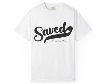 May include: White t-shirt featuring the word "Saved" in a black script font. Underneath, the text "Romans 10:9" is printed in a smaller, black font. The t-shirt is made of a soft material.