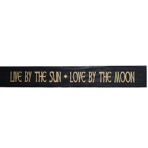 May include: Black wooden sign with gold lettering that reads "Live by the sun - Love by the moon".